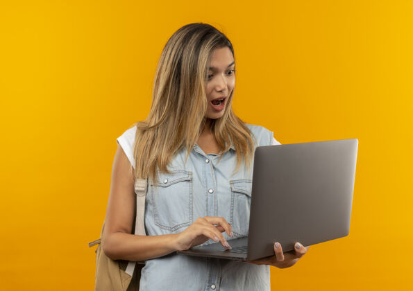使用印象深刻的年轻漂亮的女学生穿着背包使用 看着笔记本电脑孤立在橙色学生背部橙色