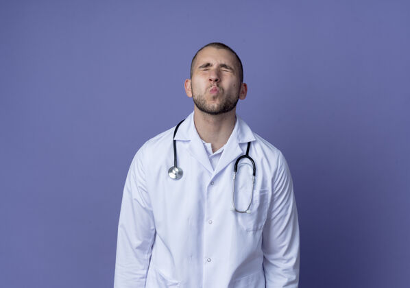 眼睛年轻的男医生穿着医用长袍 脖子上戴着听诊器 做着亲吻的动作 闭着眼睛 在紫色的背景上隔离 留有复印空间医疗戴着手势