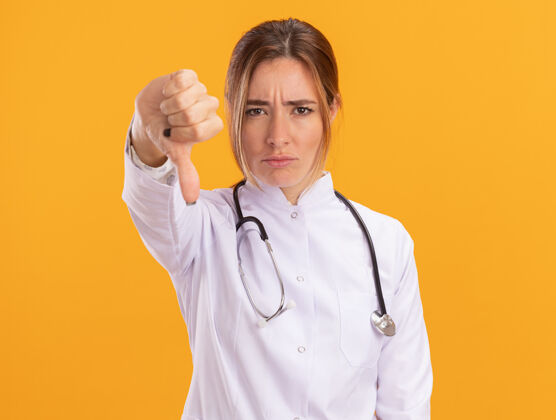 拇指年轻的女医生穿着医用长袍 听诊器显示拇指朝下 隔离在黄色的墙上穿着向下听诊器