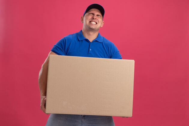 穿紧张的年轻送货员穿着制服 戴着帽子 手里拿着一个大箱子 被隔离在粉红色的墙上送货年轻盒子