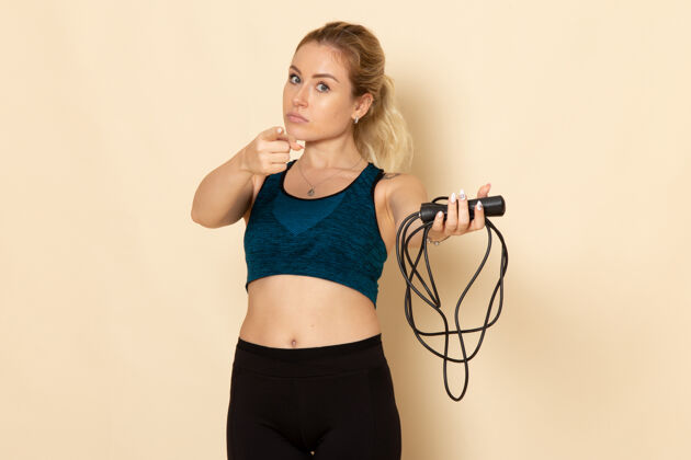 漂亮正面图身着运动装的年轻女性手持跳绳在浅白的墙上健身美容锻炼身体运动轻绳子跳过