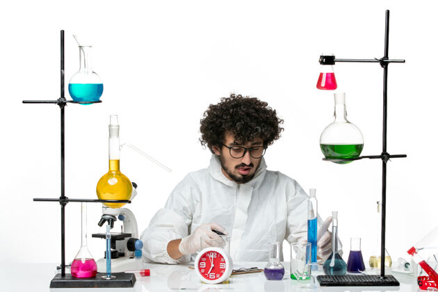 实验室正面图身穿白色特制套装的年轻男性科学家手持蓝色溶液 在白色墙上写笔记容器化学科学