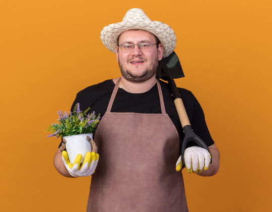 帽子微笑着的年轻男园丁戴着园艺帽和手套拿着铁锹 花盆里的花被隔离在橙色的墙上持有穿黑桃