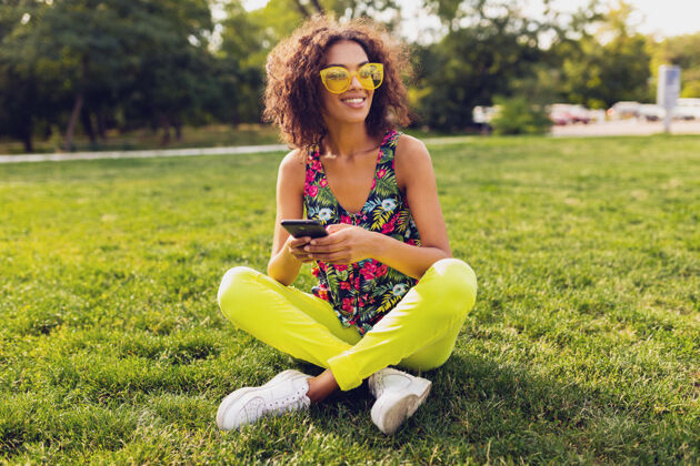 非裔美国人年轻时尚微笑的黑人女子用智能手机听音乐无线耳机在公园里玩得开心 夏日时尚多彩的风格 坐在草地上 戴着黄色太阳镜音乐手机耳机