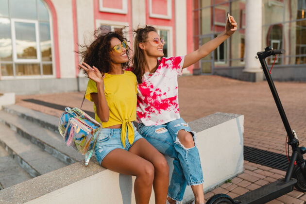 朋友快乐的年轻女孩朋友们微笑着坐在街上用手机自拍 女人们一起开心欢呼相机非洲裔