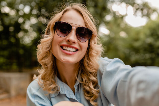 快乐迷人的金发微笑的白牙女人穿着夏装在公园里散步在电话里自拍肖像时尚春天