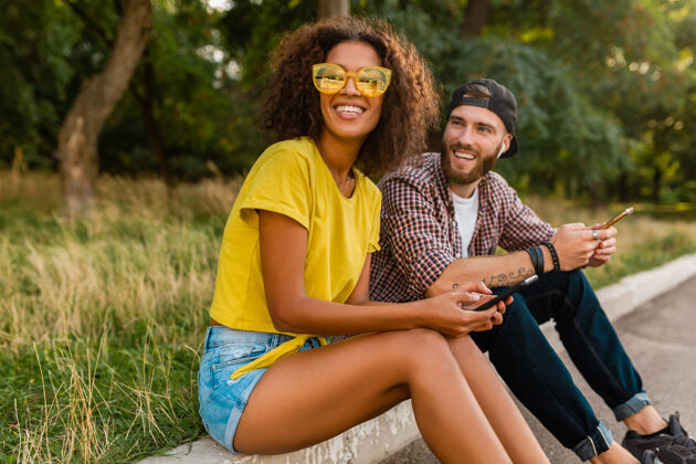 多种族快乐的年轻微笑的朋友坐在公园里用智能手机 男人和女人一起玩酷耳机兴奋