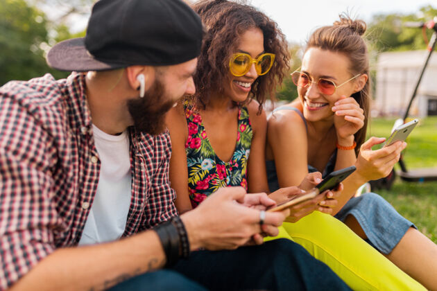 多彩快乐的年轻朋友们坐在公园里用智能手机微笑 男人和女人一起玩团体友谊女人