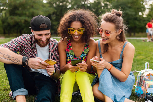 微笑快乐的年轻朋友们坐在公园里用智能手机微笑 男人和女人一起玩人兴奋多民族