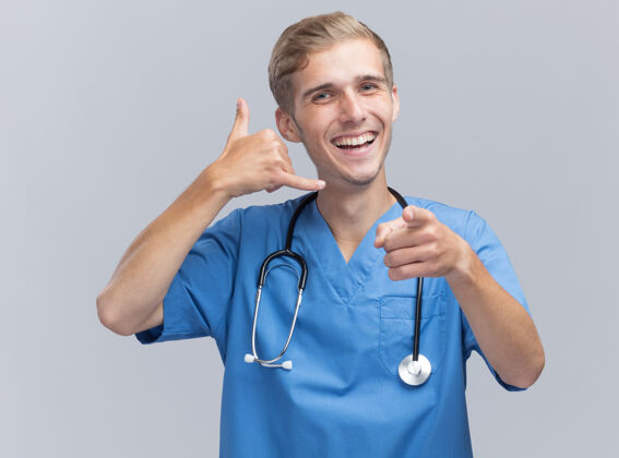 手势面带微笑的年轻男医生 穿着医生制服 听诊器显示打电话的手势 白墙上孤立着几个点微笑显示男性