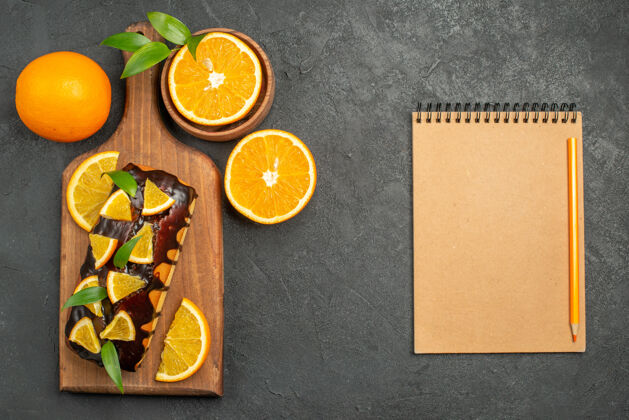 纸板上图：美味的蛋糕和切好的橙子放在砧板上 旁边的笔记本放在黑桌子上容器切割健康