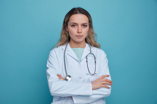 女性严肃的年轻女医生 穿着医用长袍 戴着听诊器 围着脖子 以封闭的姿势站着严重蓝色长袍