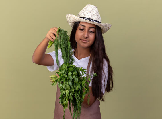 围裙快乐的年轻园丁女孩 围裙戴着夏帽 手里拿着新鲜的草药 站在绿色的墙上 脸上充满自信地微笑着帽子草药花园