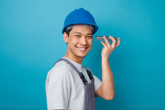 安全面带微笑的年轻建筑工人戴着安全帽 穿着制服 站在侧视图上 手持手机靠近耳朵手机微笑蓝色