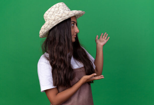 花园穿着围裙 戴着夏帽的年轻园丁姑娘站在绿色的墙上 一边看一边 手里拿着什么东西介绍围裙年轻的