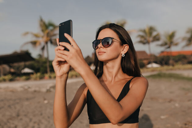 放松时尚可爱的女人戴着墨镜深色头发在沙滩上用智能手机拍照散步女人快乐