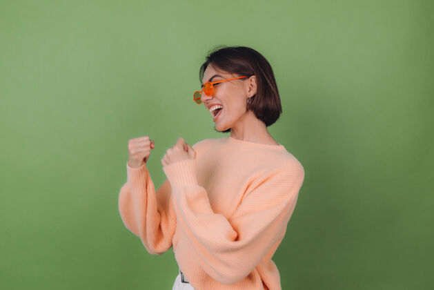 微笑年轻时尚的女士穿着休闲的桃色毛衣 戴着橙色眼镜 隔着绿橄榄色的墙壁积极地做着赢家的手势握紧拳头复制空间赢家女性欢呼