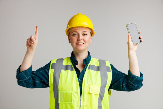 指着面带微笑的年轻女建筑工人戴着安全帽和安全背心手持手机指着上微笑工人白色