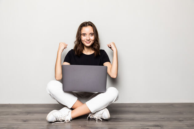 工作漂亮的女人拿着现代化的笔记本电脑坐在地板上 用胜利的姿态战胜了灰墙房子自由职业者快乐
