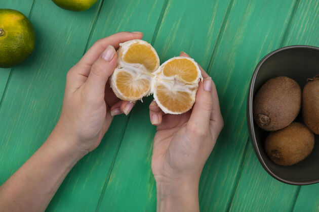 观点俯视图：一个女人手里拿着剥了皮的橘子 手里拿着碗里的猕猴桃 站在绿色的墙上持有果皮食物