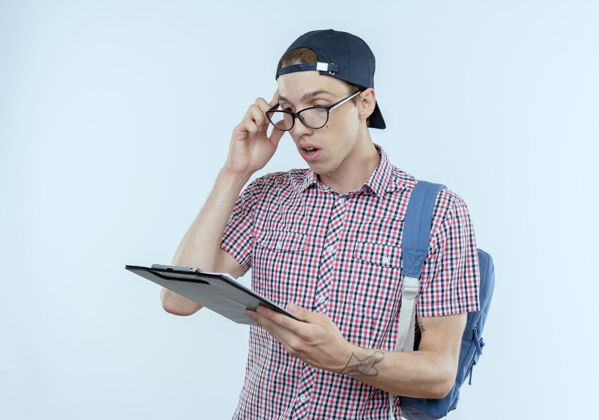 年轻人困惑的年轻学生男孩背着背包 戴着眼镜 戴着帽子 拿着剪贴板看着帽子背包剪贴板