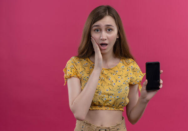 电话惊讶的年轻女孩拿着手机 把手放在脸颊上孤立的粉红色墙壁与复制空间女孩握着手机