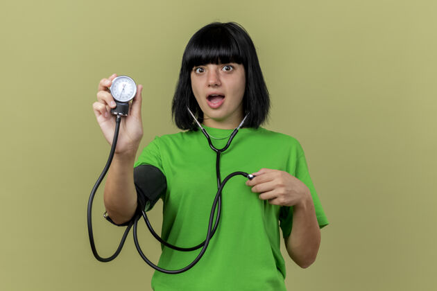 橄榄令人印象深刻的年轻病高加索女孩戴听诊器显示血压计隔离在橄榄绿的墙壁与复制空间印象听诊器血压计