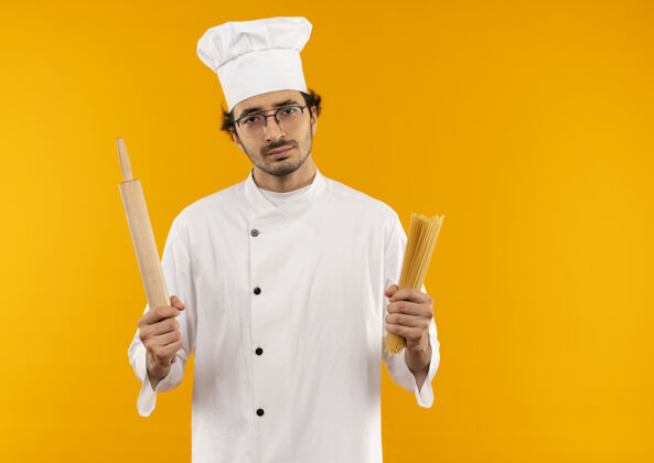 眼镜穿着厨师制服 戴着眼镜 拿着意大利面和擀面杖的年轻男厨师被隔离在黄色的墙上不愉快别针意大利面