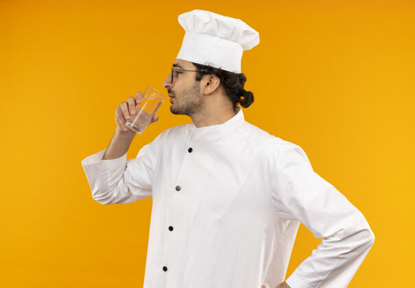 站立站在侧视图年轻的男厨师穿着厨师制服和眼镜喝水隔离在黄色的墙上年轻男性穿着