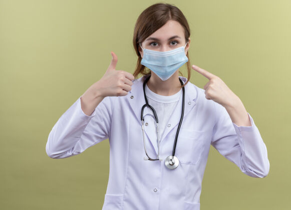 医学年轻的女医生穿着医用长袍 戴着口罩和听诊器 竖起大拇指 指着隔离的绿色墙上的口罩表演指尖年轻人