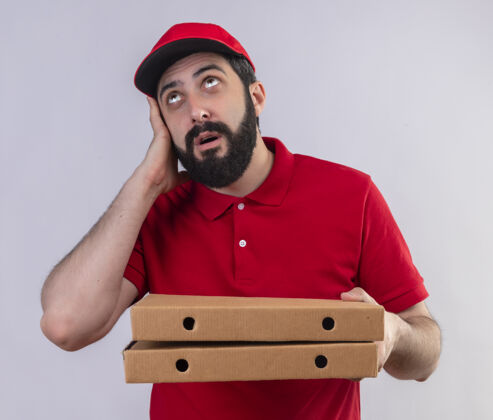年轻印象深刻的年轻英俊的白人送货员穿着红色制服 戴着帽子拿着披萨盒 手放在头上 孤立地看着白色盒子帽子手