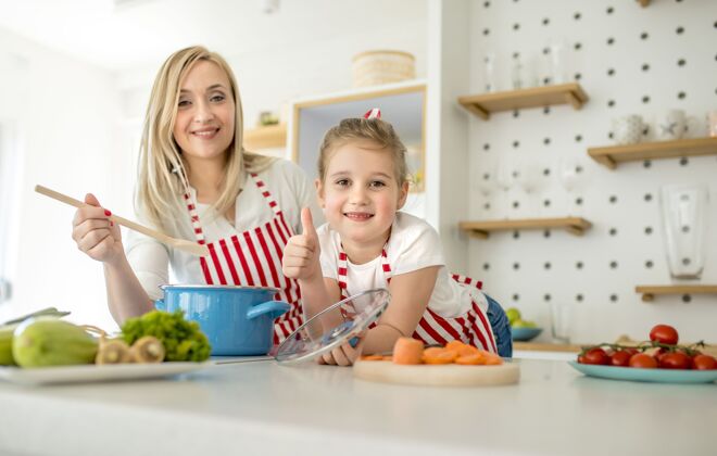 欢呼穿着相配围裙的白人母亲和女儿在厨房里微笑着摆姿势健康女儿女性