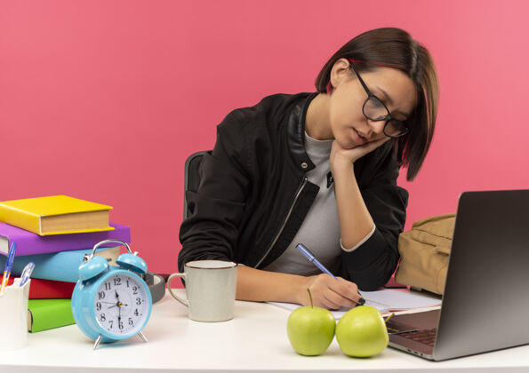 女孩专心的年轻女学生戴着眼镜坐在书桌旁用笔在记事本上写作业把手放在脸上孤立在粉色上记事本专心作业