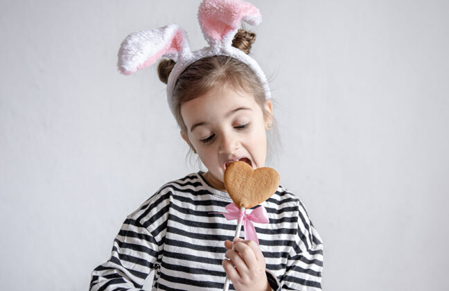兔子一个可爱的小女孩正在用棍子咬复活节姜饼 头上戴着装饰性的兔子耳朵童年咬孩子