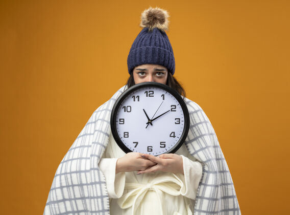 长袍虚弱的年轻白种人生病的女孩穿着长袍 戴着格子呢的冬帽 拿着时钟 从后面看相机 在橙色的背景下与世隔绝弱持有时钟