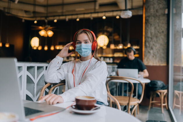 在线一个戴着耳机坐在咖啡店里的女孩冠状病毒爆发了咖啡馆面具女性