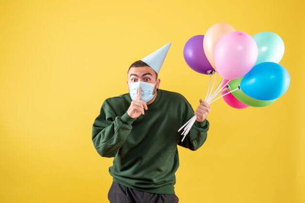 大流行正面图：黄色背景上戴着无菌面具手持彩色气球的年轻男性喜剧演员举行年