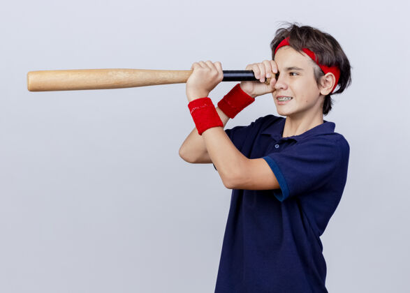 立场面带微笑的年轻帅气的运动男孩戴着头带和戴着牙套的腕带站在侧视图中拿着棒球棒用它作为望远镜隔离在白色背景上年轻背带望远镜