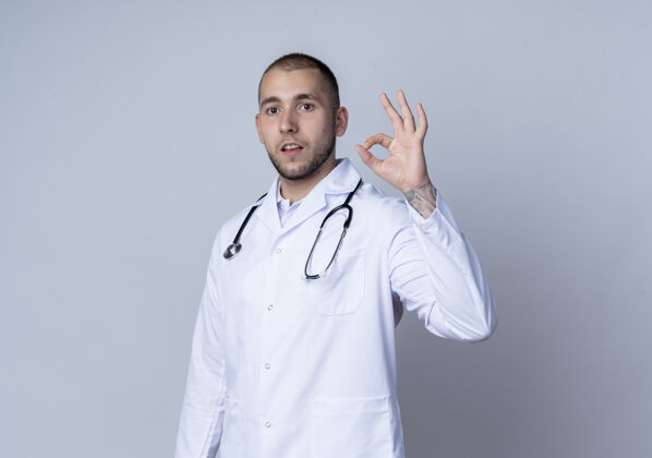 男性自信的年轻男性医生 穿着医用长袍 脖子上戴着听诊器 做着ok的手势 隔离在白色的空白处标志副本年轻