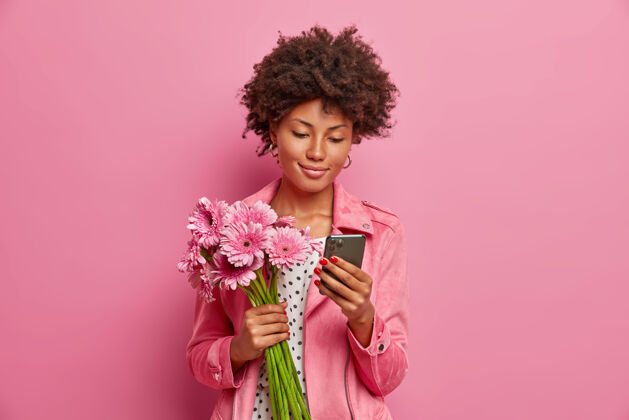 风格高兴的年轻女子有非洲头发 得到非洲菊花束作为礼物 摆出美丽的鲜花和手中的智能手机 在网上发送信息 得到惊喜的礼物休闲设备非洲菊