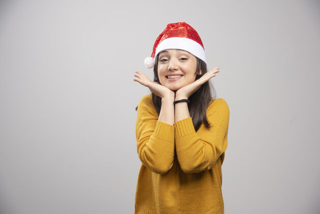 模型戴着圣诞帽的漂亮女人在灰色的墙上感到快乐女孩圣诞老人人
