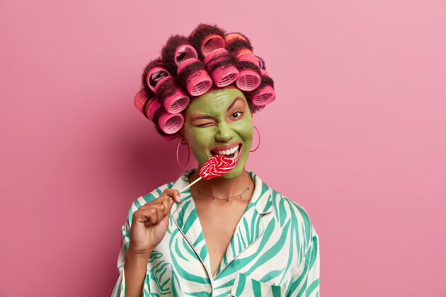 棒棒糖快乐的年轻非洲裔美国女人眨眼 咬美味的棒棒糖 脸上涂绿色面膜 卷发器 穿着随意 接受美容治疗皮肤护理面具微笑