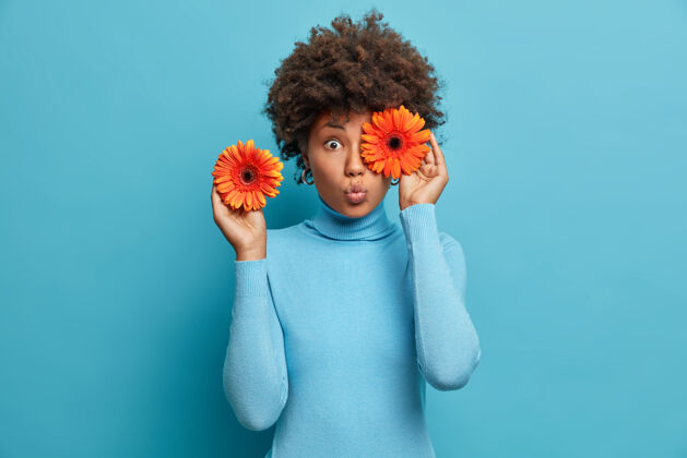 女性一个非常惊讶的女人用橙色非洲菊遮住眼睛 拿着鲜花 为特殊场合装饰大厅 穿着蓝色高领毛衣 站在室内美国高兴高兴
