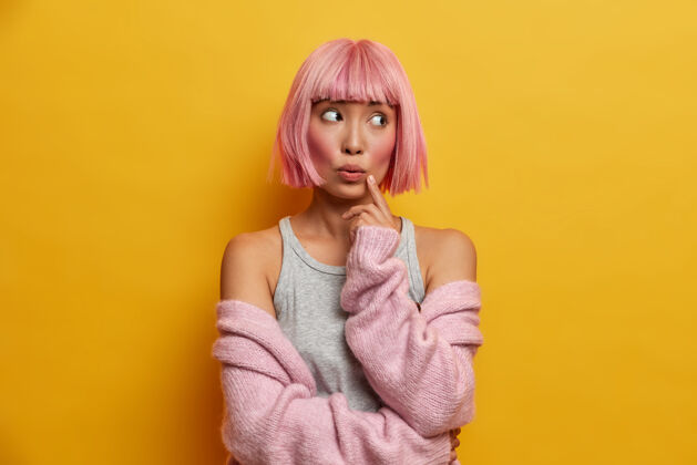 困惑年轻体贴惊讶的亚洲女人的画像 玫瑰色的头发 手指贴在脸上 脸上露出疑惑 穿着温暖的针织毛衣粉色年轻粉色头发