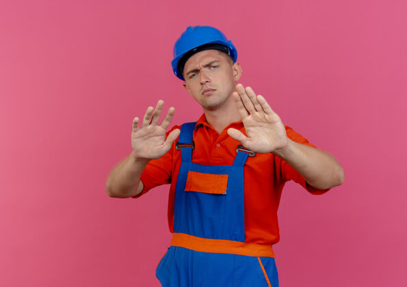 严格严格的年轻男性建设者穿着制服和安全帽显示停止手势粉红色头盔年轻男性