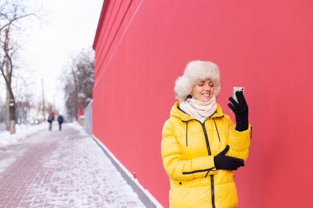脸在一个冬天阳光明媚的日子里 一个穿着暖和衣服的红墙背景上的快乐的年轻女子在雪城的人行道上微笑着打电话广场说话手机