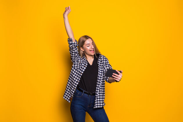 手机一个快乐的年轻女子手持手机 独自站在黄色墙壁上庆祝的画像魅力使用电话