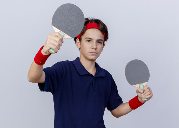 伸展自信的年轻帅气的运动男孩戴着头带和护腕 戴着牙套 看着前方 拿着乒乓球拍 向前方伸展 隔离在白色的墙壁上人表情抱着