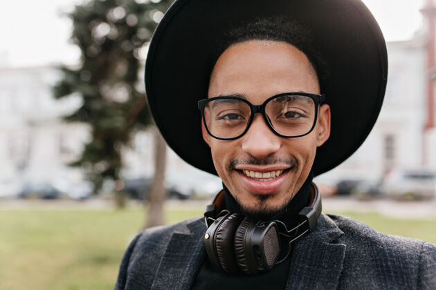 帅气幸福的非洲男模的特写照片 黑眼睛站在模糊的大自然上戴着黑帽子和耳机的时髦男人走在街上的户外肖像户外年轻年轻