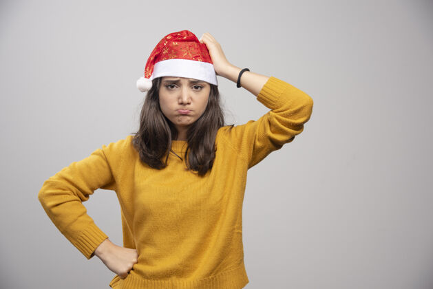 姿势一个戴着圣诞老人红帽子在白墙上摆姿势的心烦意乱的女人年轻墙圣诞老人
